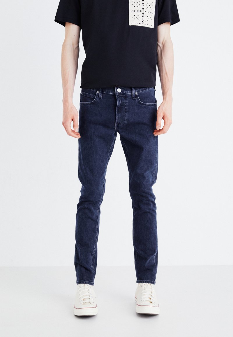 Jeans bleu foncé au design slim, avec une taille standard et un style classique à cinq poches. Portés avec un t-shirt noir.