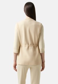 Beige Strickweste mit geripptem Kragen und Bündchen, tailliert mit einem Gürtel, aus einem weichen Strickmaterial, kombiniert mit cremefarbenen Hosen.