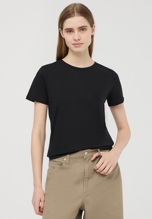 T-shirt nera a maniche corte in cotone con scollo rotondo; design aderente. Indossata con pantaloni beige chiaro, con dettagli di bottoni e tasche.