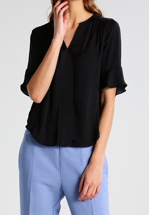 Vrouw draagt een losse zwarte blouse met ruches korte mouwen en lichtblauwe hoge taille broek, hand bij het gezicht en aan de zijkant.