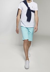 Maglietta bianca, pantaloni corti azzurri chiari e sneakers bianche. Un maglione scuro è annodato intorno alle spalle. Modello che cammina su uno sfondo neutro.