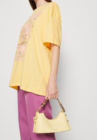 Camiseta amarilla de gran tamaño con estampado floral y acentos en rosa, combinada con un pequeño bolso amarillo con relieve de croc que cuenta con una correa de cadena.