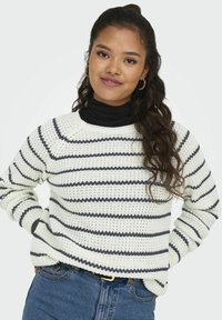 Crocheted sweater in white with navy stripes. Features a crew neckline and long sleeves, paired with a fitted black turtleneck underneath.
