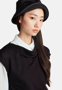 Černý pletený svetr s rolákem, nošený přes bílou košilovou košili. Modelka má také na sobě černý klobouk typu bucket. Dlouhé tmavé vlasy jí splývají dolů.