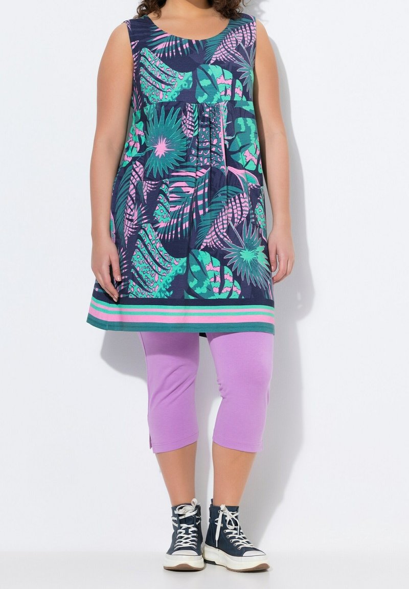 Femme portant une tunique sans manches à imprimé tropical avec des feuilles vertes et roses, des leggings capri lilas et des baskets montantes noires avec des lacets blancs.