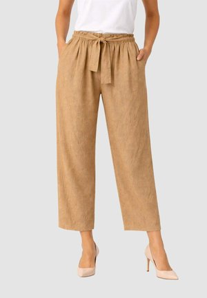 Donna con pantaloni beige a gamba larga con cintura annodata e tasche, abbinati a décolleté color nude e top bianco, in piedi davanti a uno sfondo semplice.