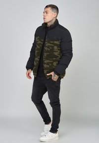 Pufová bunda s čiernym nylonovým a zeleným maskáčovým fleece, vysoký golier, zips, nosená s čiernymi džínsami a bielymi teniskami.