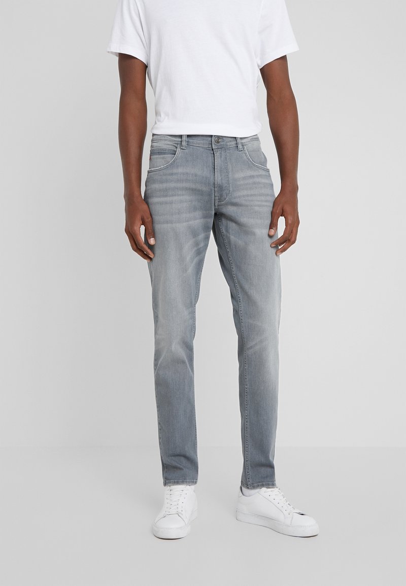 Jeans de mezclilla gris con un diseño ajustado, sutiles desgastes, cinco bolsillos y herrajes metálicos. Combinados con una camiseta blanca sencilla y zapatillas blancas.