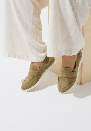 Pieds portant des mocassins en daim vert olive à enfiler avec des semelles blanches, associés à un pantalon ample en lin crème, reposant sur un bloc de bois clair.