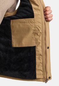 Dos manos sosteniendo una chaqueta clara abierta que muestra un forro de felpa negra y un bolsillo interior rectangular beige con costuras reforzadas.