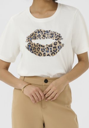 Frau trägt ein weißes Kurzarmshirt mit großem Leopardenlippen-Design, beigen Hosen und ein goldenes Armband am Handgelenk.