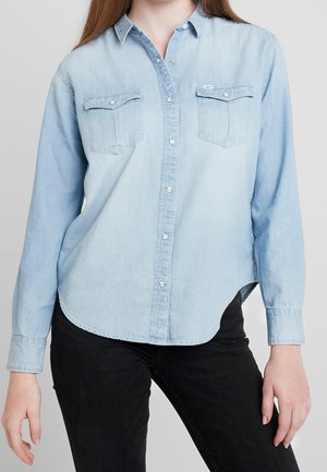 Žena v světle modré džínové košili s knoflíky a dvěma náprsními kapsami, černých kalhotách, stojící před neutrálním pozadím.