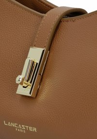 Bruine leren handtas met een gestructureerde textuur, voorzien van een gouden sluiting en logo met een gladde afwerking. Het ontwerp heeft een ronde vorm en een elegante handgreep.