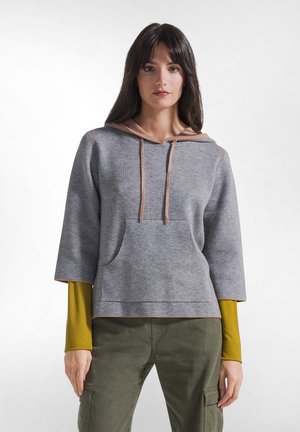 Femme portant un sweat à capuche gris à manches courtes superposé sur une chemise moutarde à manches longues et un pantalon cargo vert olive, debout devant un fond uni.