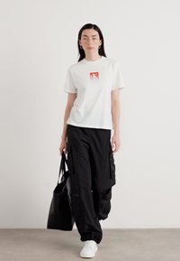 Witte katoenen t-shirt met een rood CK-logo, gecombineerd met losse zwarte cargobroeken en witte sneakers, met een zwarte tas. Neutrale achtergrond.