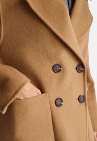 Kamelově zbarvený vlněný kabát s dvouřadým zapínáním, velkými tmavými knoflíky a boční kapsou. Tkanina má jemnou texturu.