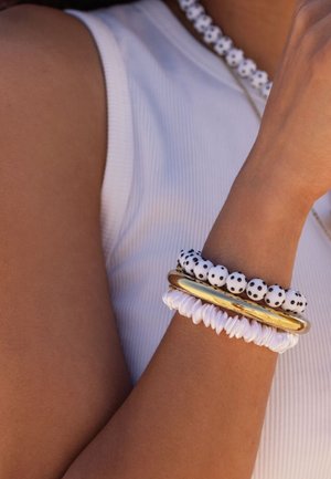 Bras portant un débardeur blanc avec trois bracelets : perles blanches à pois noirs, jonc doré lisse et bande blanche texturée.