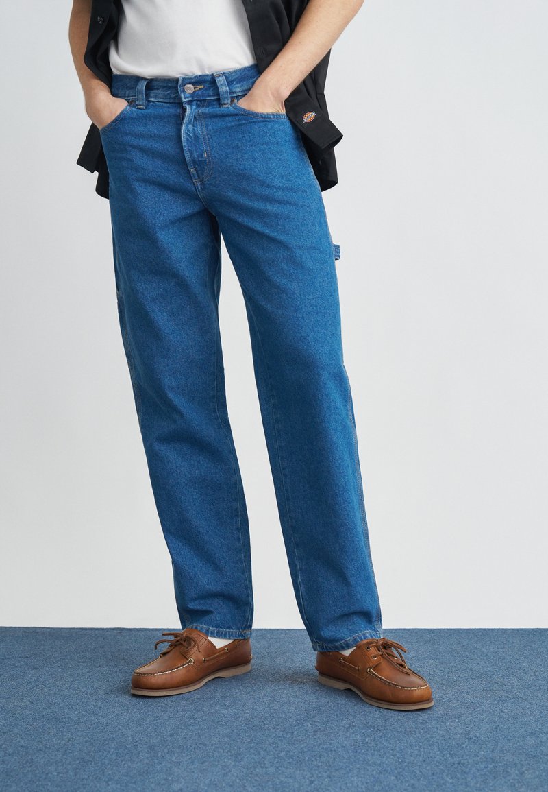 Jean en denim bleu à coupe droite avec un léger effet délavé. Associé à des chaussures en cuir marron. L'arrière-plan est gris clair avec un tapis bleu.