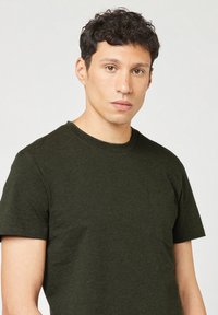 Donkergroene t-shirt met korte mouwen en een ronde hals, voorzien van een borstzakje. De stof heeft een zachte, textuele uitstraling. Minimalistisch ontwerp.