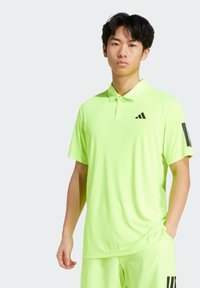 Világos zöld pólóing klasszikus gallérral, rövid ujjakkal, háromgombos felsőrészen és fekete adidas logo-val a mellkasán és az ujján.