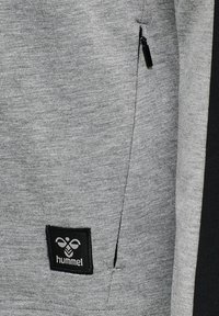 Szary sweatshirt z boczną kieszenią na zamek, wykonany z textured materiału. Czarna naszywka z logo na dole z marką "hummel".