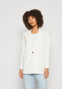 Bílý blazer s jedním hnědým knoflíkem, klopami a dvěma předními kapsami, kombinovaný s bílým topem a světlemodrými džíny.