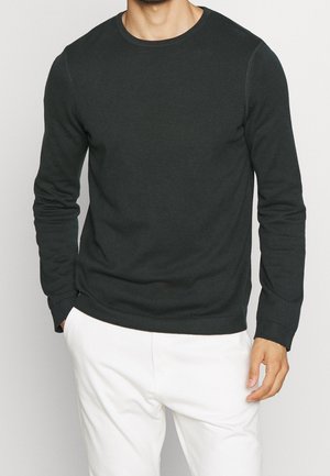 Homme portant un pull vert foncé à manches longues et col rond, et un pantalon blanc, une main dans la poche, sur un fond clair uni.