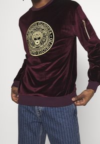 Felpa crewneck in velluto bordeaux con emblema di un leone dorato e testo "Glorious Gangsta". Polsini e vita a coste; dettaglio con tasca con zip.