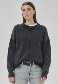 A model wears a dark oversized sweater with a ribbed collar, paired with light jeans and a black belt, set against a neutral background.