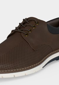 Chaussure marron en cuir à lacets avec détails perforés, œillets dorés et semelle en caoutchouc blanche avec accents noirs sur la bande de roulement.