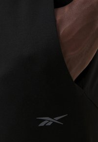 Svart ermeløs topp laget av glatt stoff, med en subtil tekstur og et reflekterende logo på nedre venstre side.