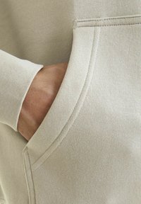 Beige sweatshirt nærbillede, der viser en glat stofstruktur, ribbet manchet og en sidelomme med synlige syninger.