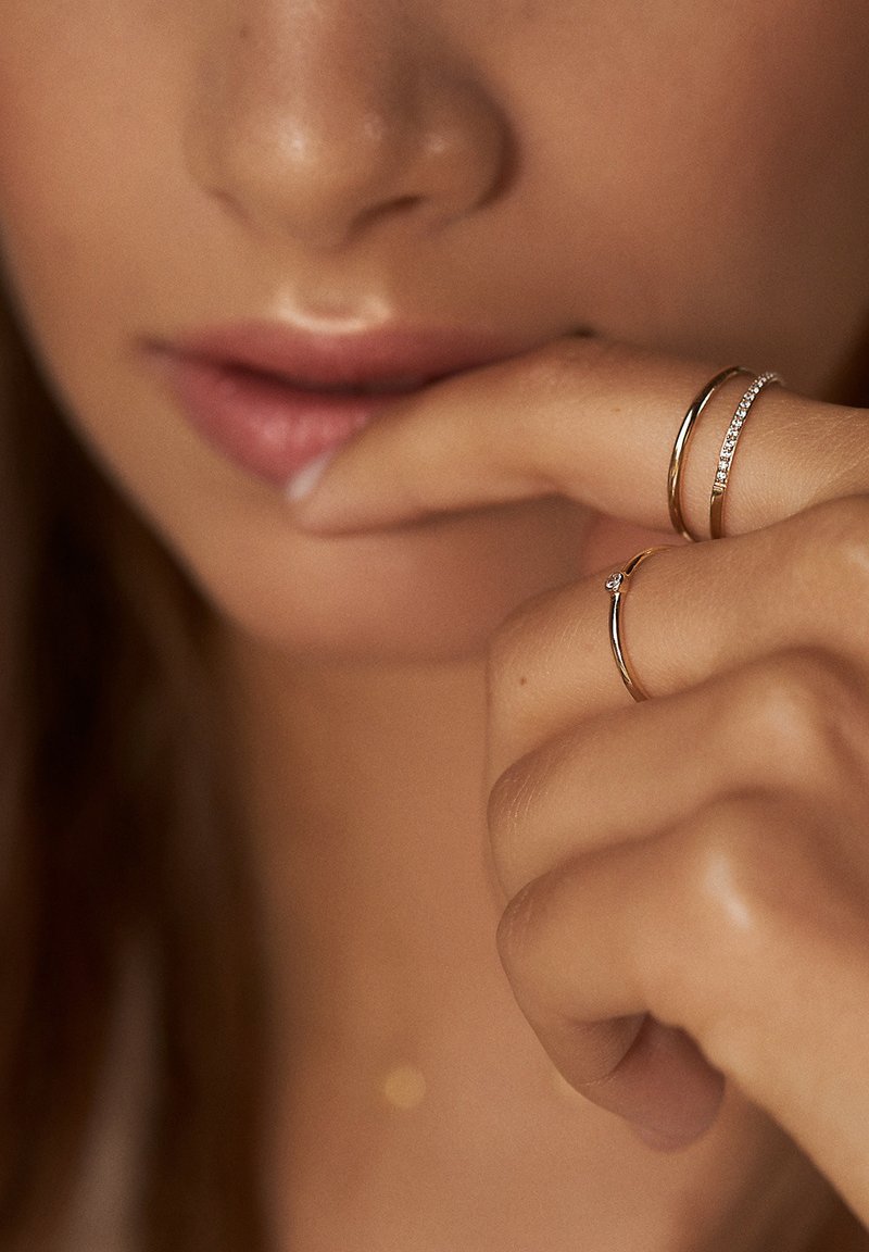 Trois anneaux sur une main : une fine bande en or, une bande en or avec un petit diamant, et une bande en or ornée d'une rangée de petites pierres, toutes brillantes.