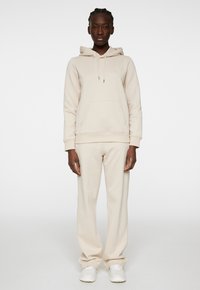 Felpa beige con tasca frontale e cordino, abbinata a pantaloni della tuta tapered coordinati, caratterizzati da una texture liscia e una vestibilità casual.