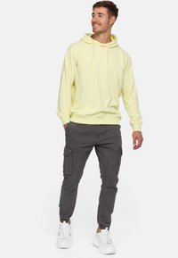Sudadera amarilla clara con bolsillo tipo canguro y capucha con cordón, combinada con pantalones cargo gris oscuro que tienen bolsillos laterales y tobillos con puños.