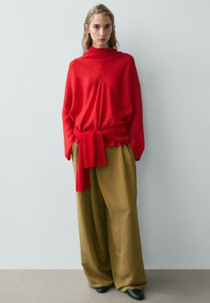 Donna che indossa un maglione a collo alto rosso largo con cintura abbinata annodata in vita, pantaloni larghi verde oliva e scarpe nere, in piedi contro un muro.