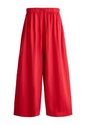 Femeie cu păr blond ondulat, purtând un tricou alb cu text, pantaloni roșii largi și sandale negre cu platformă, stând în picioare pe un fundal simplu.