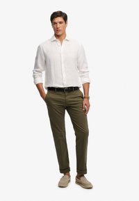 Hombre con camisa blanca de manga larga, pantalones verde oliva remangados, zapatos beige sin cordones, cinturón negro y reloj de pulsera, de pie con una mano en el bolsillo.