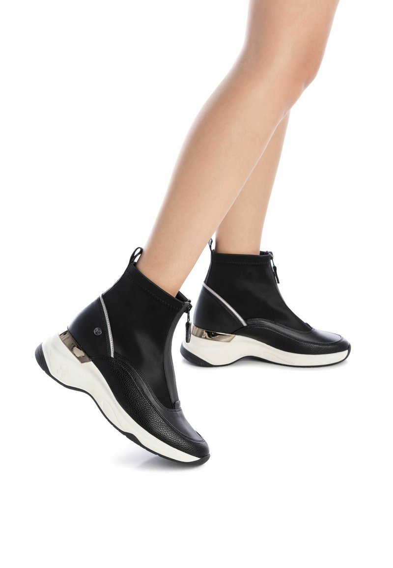 Bottines noires avec un dessus lisse, présentant un revêtement texturé, une fermeture éclair, et une semelle blanche contrastante avec des accents métalliques.