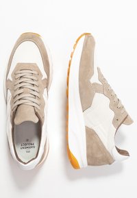 Pari beige ja valkoisia lenkkikenkiä oransseilla pohjilla, verkkokudosta ja mokkapintaa, sekä nauhoja, esitelty ylhäältä ja sivulta valkoista taustaa vasten.