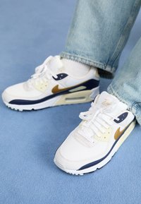 Vita atletiska skor med navy och guld accenter, mesh- och syntetmaterial, med en texturerad sula och snörningsdesign.