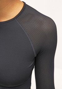 Närbild av skuldra och överarm som bär en åtsittande svart långärmad topp med nätmaterial på skulderpartiet och ärmen.