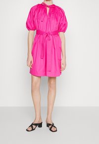 Woman wearing a bright pink short dress with puffed sleeves, a tied neckline, a matching belt, and black heeled sandals.