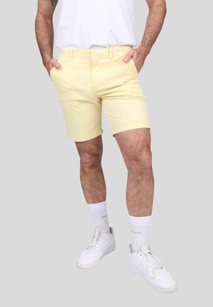 Shorts en coton jaunes avec une coupe régulière, devant plat, poches latérales et un tissu légèrement texturé, portés avec des sneakers blanches.