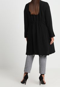 Černý kabát po kolena s úzkým pasem, dlouhými rukávy a bočními kapsami, v kombinaci se šedými károvanými kalhotami a černými lodičkami na vysokém podpatku.