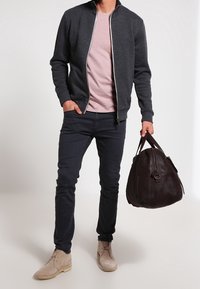 Hombre con chaqueta oscura de cremallera, camiseta rosa, jeans negros, botas beige, sosteniendo un gran bolso de cuero marrón oscuro por las asas.