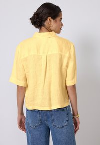 Camicia gialla in lino a maniche corte con una vestibilità rilassata, dettaglio sul giromanica e orlo dritto. Indossata con jeans in denim blu.