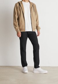 Beiger Kapuzenpullover mit Reißverschluss und Kordeln über einem weißen T-Shirt, kombiniert mit schwarzen Hosen und weißen Turnschuhen, stehend auf einem neutralen Bodenhintergrund.