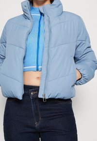 Svetle modrá péřová bunda s vysokým límcem a zipovým uzávěrem, se vzorem horizontálního prošívání a jemně hladkým povrchem.