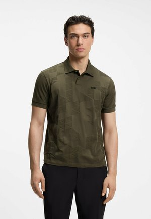 Mann trägt ein olivgrünes, gemustertes Poloshirt mit kurzen Ärmeln und schwarze Hose, schaut nach vorne mit neutralem Gesichtsausdruck.